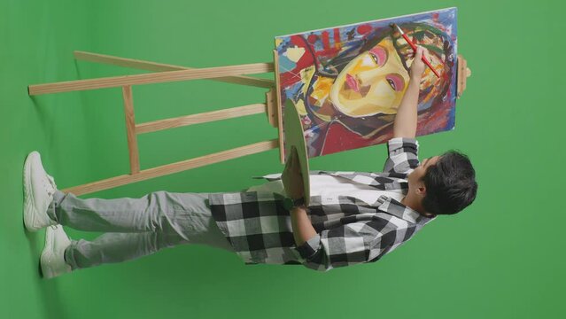 Full Body Side View Of Asian Male Artist Is Concentrated Painting A Girl On Canvas By Oil Paints And Brush In The Green Screen Studio
