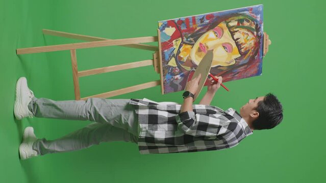 Full Body Side View Of Asian Male Artist Using Paint Brush Measuring Girl'S Face On Canvas By Oil Paints And Brush In The Green Screen Studio
