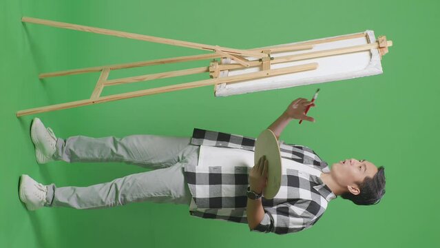 Full Body Of Asian Male Artist Crossing His Arms And Thinking Over Picture Concept While Painting On Canvas By Oil Paints And Brush In The Green Screen Studio
