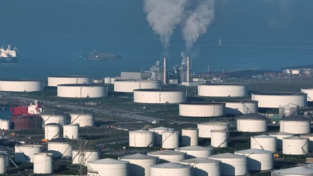 Wide aerial view of large capacity oil storage tanks near the ocean with ships in the background and industrial facility with stacks emitting steam
