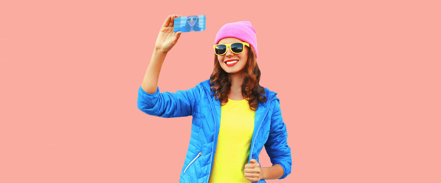 Portrait of modern happy smiling young woman taking selfie with smartphone wearing colorful hat, blue jacket on pink background