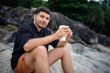 Attractive young man holding a disposable glass with coffee drink on the rocks of a tropical beach in the morning and looking at the camera.