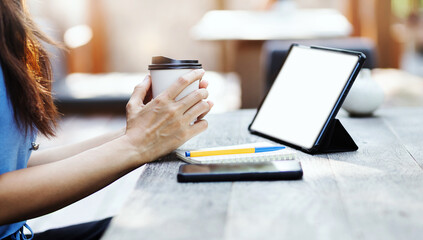 Asian freelance woman holding cup of hot coffee and working on tablet computer on wooden table at outdoor cafe. Entrepreneur woman working for her new business concept.