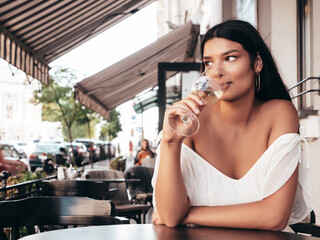 Young beautiful smiling hipster female in white dress. Sexy carefree woman sitting at cafe in the street. Positive model drinking white wine. Cheerful and happy. Outdoors at sunny day