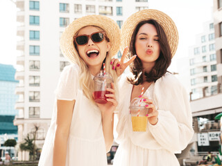 Two young beautiful smiling hipster female in trendy summer white dress and straw hats. Sexy carefree women holding and drinking fresh vegetable cocktail smoothie drink in plastic cup with straw
