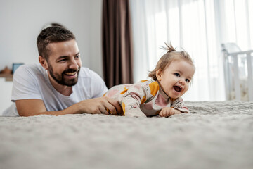 Toddler is playing with her father in a bedroom. She is crawling on a bed while father is tickling...