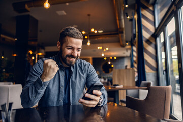 A young successful man is celebrating victory while sitting in coffee shop and looking at the phone.