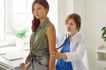 Obraz premium Teen brunette girl on checkup appointment with young woman doctor examining her in clinic. Pediatrician listens her lungs with a phonendoscope. Healthcare, child pediatrician, medicine concept.