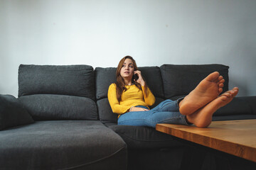 Fototapeta premium woman with brown hair and yellow shirt, wearing jeans, sitting in an armchair using a mobile phone.