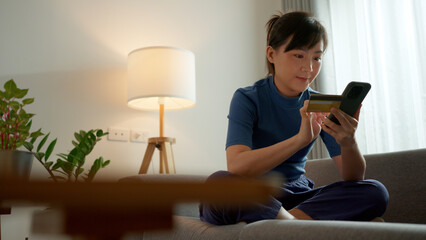 Asian woman sitting on sofa using smart phone and credit card for shopping online at home.