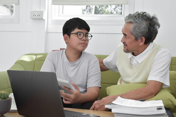 Obraz na płótnie Canvas Asian senior grandpa and his son are spending times together at home to connect headphone with his laptop to watch and to play online games, soft and selective focus, raising teens concept.