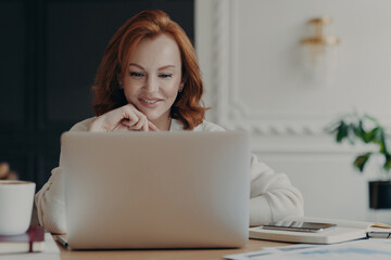 Fototapeta premium Experienced red haired businesswoman reads publication on modern laptop computer, concentrated at screen with pleasant smile, makes research for planning startup, checks mail box, works from home