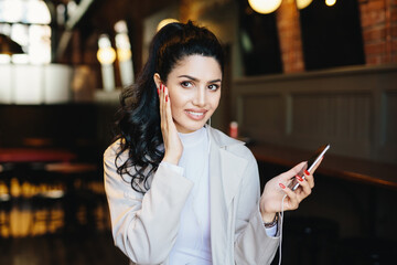 Brunette young model with lovely appearance having delicate hands dressed in white jacket listening to her favorite tracks on earphones, using online music app on cell phone at cozy restaurant