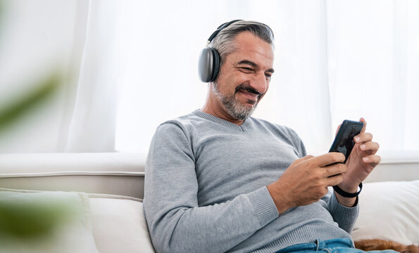 Portrait Of Senior Old Caucasian Man Stay Home Sit On The Sofa  Living Room Listen To Music Head Phone With Lovely Dog. Happy Moment Of Pure Love Middle Age Male Playing With Little Dog In Winter