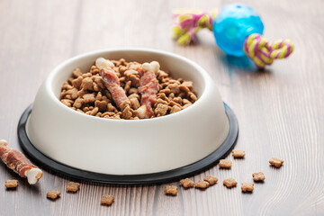 A bowl with dog food, dog treats and toys on a wooden floor.
