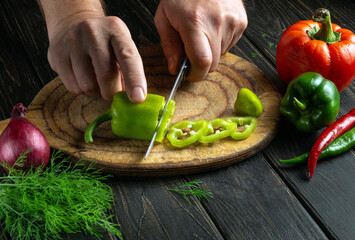 The hands of a cook with a knife cut green peppers on a cutting board to prepare a vitamin salad....