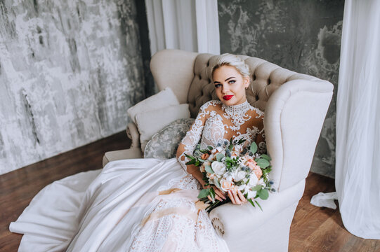 A beautiful, young blonde bride in a long lace dress sits on a sofa against a gray wall in the studio, interior. Wedding photography, portrait.