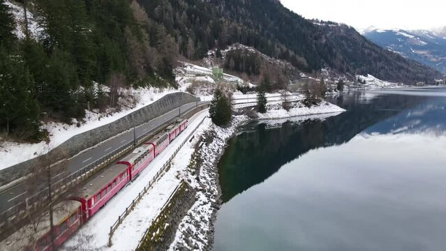 Dark green lake in the heart of Swiss Alps in wintertime, famous red glacier express train passing through the snowy mountains railway, aerial view of amazing landscape in Switzerland, travel