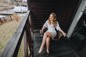 A beautiful curly blonde bride in lingerie, a dressing gown sits on a wooden chair on the veranda with a glass of champagne in her hands. Wedding photography, portrait.