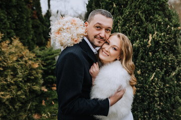 A stylish bearded groom in a black coat and a beautiful smiling blonde bride in a white dress, fur coat with a bouquet of reeds gently hug outdoors in the park. Wedding photography, portrait.