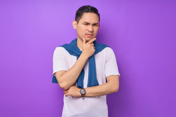 Obraz premium Pensive young handsome Asian man wearing white t-shirt touching her chin, thinking and lost in thought while looking at the camera isolated on purple background. People emotions lifestyle concept