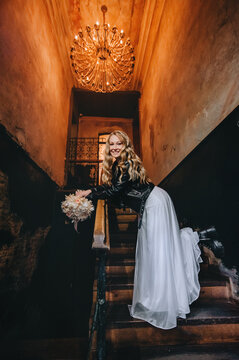 A beautiful cool bride, biker, rock lover in a black leather jacket, white dress with a bouquet stands on the steps indoors against the background of a wall with a chandelier. wedding photography.