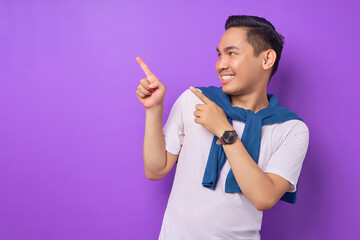 Smiling young Asian man wearing white t-shirt pointing fingers aside at blank space for advertising text isolated over purple background. people lifestyle concept