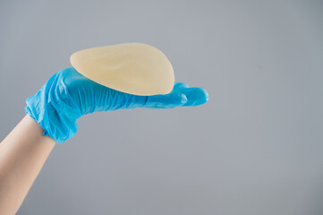 A female hand in gloves holds a teardrop-shaped breast implant on a gray background. 
