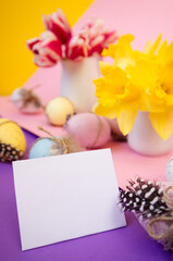 White empty postcard near decorating easter eggs and spring flowers on colored background