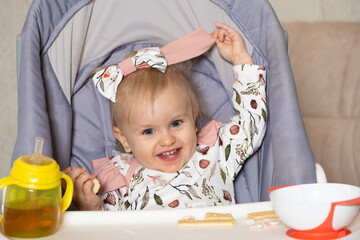 Naklejka premium A happy little girl sits in a high chair with a bottle and a plate and looks into the camera smiling.