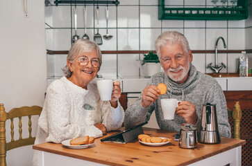 Portrait of attractive smiling senior couple having breakfast together sitting at kitchen table. Elderly 70s husband and wife enjoying relaxed retirement looking at camera