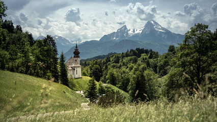 Fototapeta na wymiar Kirche zwischen grünen Bäumen und Bergpanorama im Hintergrund. In Berchtesgaden mit Watzmann im Sommer.