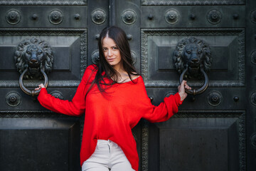 Gorgeous young brunette woman in red sweater and white jeans posing against antique metal door, holding decorative door handles executed in a lion head form. Vogue  and woman lifestyle concept.