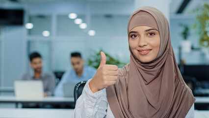 Islam Muslim businesswoman in hijab ethnic happy successful woman office worker entrepreneur worker manager company CEO looking at camera showing thumb up good fine agree approve hand gesture indoors