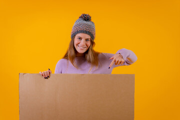 Portrait of a woman in a wool cap pointing at a cardboard sign on a yellow background