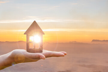Naklejka premium hand holds wooden flat house against golden sunset background. protection or property concept.
