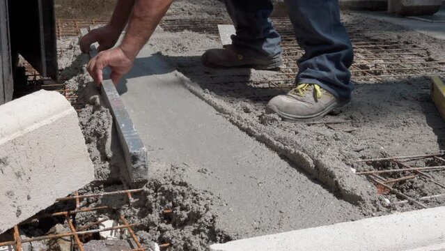 Professional building contractor in dirty clothes and boots using metal levelling tool for aligning or smoothing fresh cement surface on construction site, process of reinforcing basement mesh with