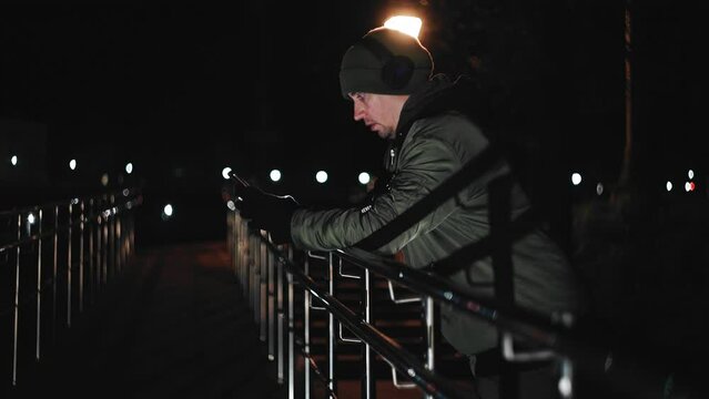 At night in the park, a guy leans on the handrails and looks at the phone screen. he has headphones on his head