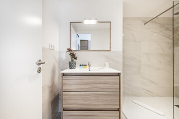 Fototapeta na wymiar Bathroom with wooden cabinet of drawers with white porcelain sink, mirror with matching frame, marble tiles and shower cabin with glass screen