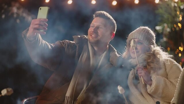 multi ethnic friends taking selfie in picnic in winter evening, roasting marshmallows and have fun