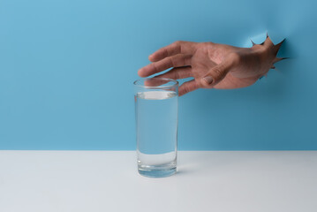 Male hand with glass of water breaks through blue paper background.