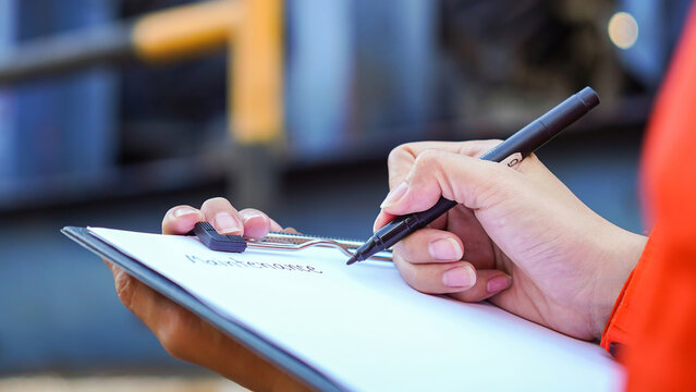 An engineer is writing on paper the to the maintenance detail with a heavy pumping machine as blurred background. Industrial expertise occupation working scene, selective focus at hand's part.
