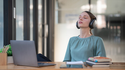 Young woman enjoying listening to music wearing headphones feeling relaxed at an office desk.