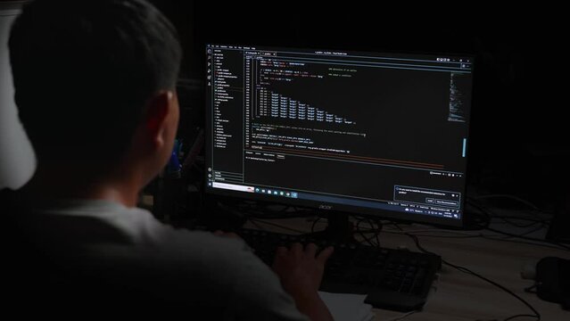 Back view young asian man sitting at desk using computer programming debugging improving binary algorithm code in workstation By focusing on the monitor and blurring on people  in the dark