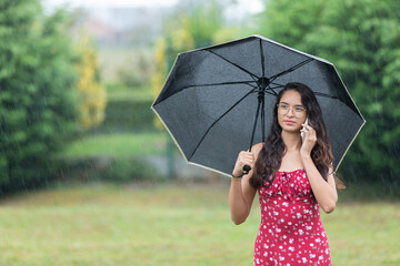 Ethnic woman with umbrella having phone argument in park on rainy day 