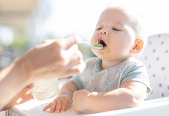 Mother spoon feeding her baby boy child in baby chair with fruit puree on a porch on summer...