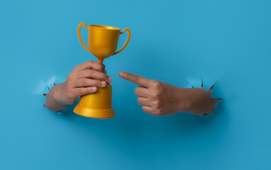 Female hand holding a champion golden trophy breaks through blue paper background.