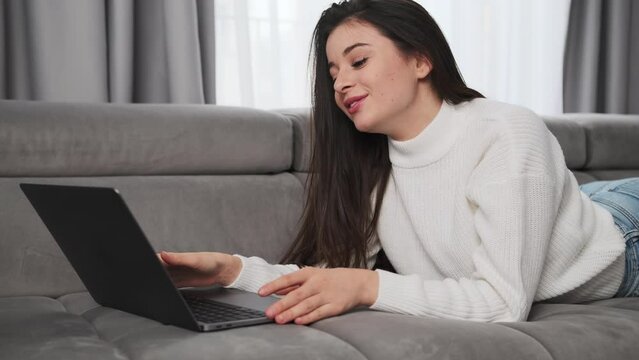 Cheerful woman talks with parents on video call via Notebook lying on sofa in living room. Brunette lady uses laptop for online communication
