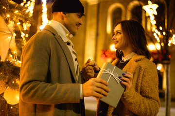 Stylish couple holding hands and gift near Christmas tree on street in evening 