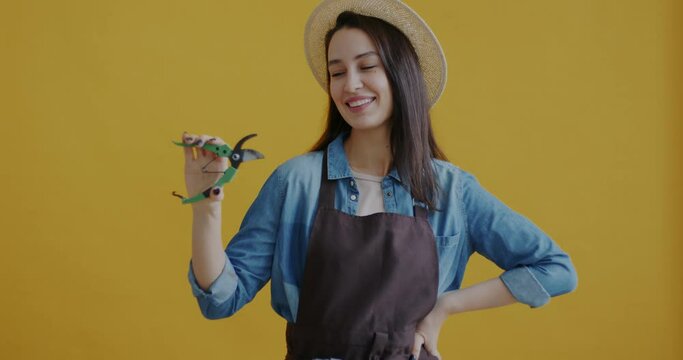 Potrait of young woman gardener holding pruning scissors and smiling standing on yellow background. Gardening tools and positive emotion concept.